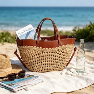 Op de afbeelding: Een tote bag met een bruin lederen bovengedeelte met witte stiksels en een geweven beige strooien onderkant. De tas heeft twee lederen handvatten en een afneembare schouderriem. Een tijdschrift zit in de open bovenkant van de tas gestoken.
