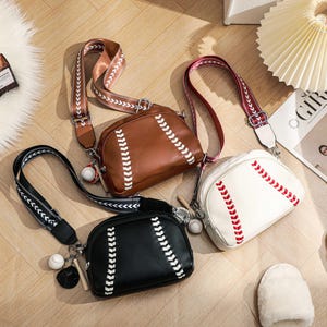 Puede incluir: Tres bolsos cruzados con temática de béisbol en marrón, blanco y negro. Cada bolso presenta costuras blancas, una correa estilo béisbol y un dije de béisbol. Los bolsos están sobre una superficie de madera.