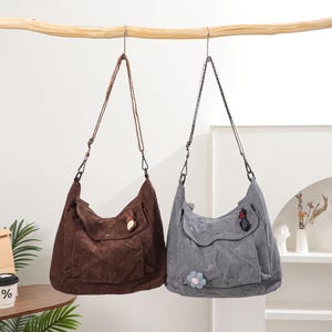 May include: Two corduroy shoulder bags hanging from a wooden branch. One is brown, the other is gray. Both have a large front pocket and adjustable straps. The brown bag has a small, light-colored accent, while the gray bag has a flower and a red accent.
