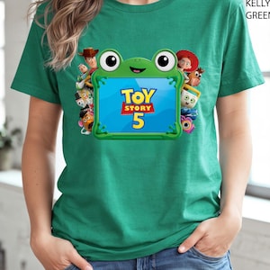 May include: A kelly green short-sleeve t-shirt featuring a graphic of a green frog-shaped frame surrounding the text Toy Story 5. The frame is decorated with various animated character figures including a cowboy, a cowgirl, and a potato-shaped toy.
