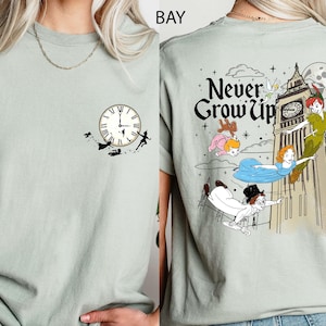 May include: A light green t-shirt featuring a clock design with silhouettes of Peter Pan and Tinkerbell on the front. The back of the shirt has a graphic of Peter Pan, Wendy, and other characters with the text "Never Grow Up".