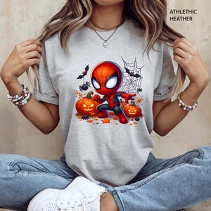 May include: Athletic heather t-shirt featuring a cartoon Spider-Man character with pumpkins, bats, and spiderwebs. The design has a Halloween theme with orange and black accents. The text "ATHLETIC HEATHER" is printed at the top right.