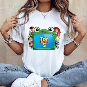 May include: A white short-sleeved t-shirt featuring a graphic of a green frog-shaped tablet frame. The screen displays the text Toy Story 5 in yellow and red. Surrounding the frog are various animated characters including a cowboy and a cowgirl.