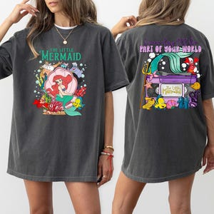 May include: Dark gray t-shirt featuring a colorful graphic of Ariel from The Little Mermaid on the front, with the text "THE LITTLE MERMAID". The back of the shirt has a graphic with the text "Someday I'll be part of your world".