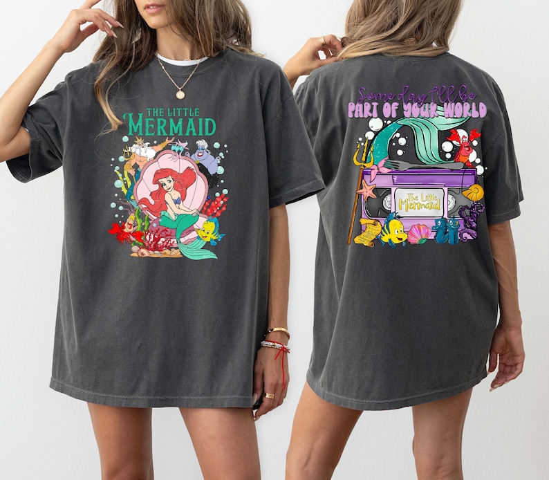 May include: Dark gray t-shirt featuring a colorful graphic of Ariel from The Little Mermaid on the front, with the text "THE LITTLE MERMAID". The back of the shirt has a graphic with the text "Someday I'll be part of your world".
