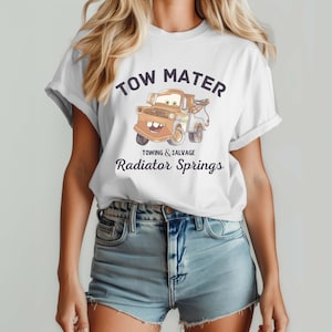 May include: White t-shirt featuring a cartoon image of Tow Mater, a tow truck character, with the text "TOW MATER" above the image and "TOWING & SALVAGE Radiator Springs" below. The shirt is worn by a person.