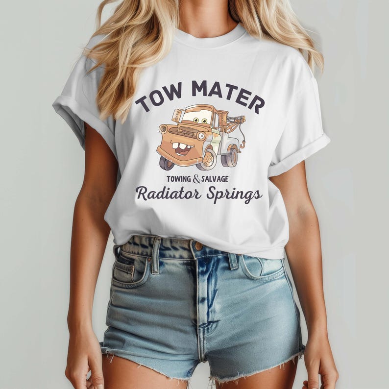 May include: White t-shirt featuring a cartoon image of Tow Mater, a tow truck character, with the text "TOW MATER" above the image and "TOWING & SALVAGE Radiator Springs" below. The shirt is worn by a person.