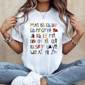 May include: White t-shirt with a colourful alphabet design featuring Disney characters. Each letter is represented by a different character, including Ariel, Belle, and Cinderella. The shirt is a casual, everyday wear item.