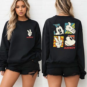 May include: Black sweatshirt featuring a Mickey Mouse design. The front displays a peace sign with the word "Mickey" in red. The back showcases Mickey Mouse's face, hand, and peace sign in a colorful, square pattern. A casual, comfortable top.