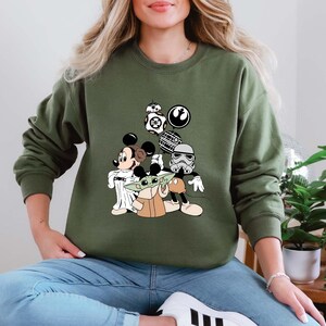 Puede incluir: Sudadera verde oliva con un dise&ntilde;o de Mickey Mouse como Jedi, Baby Yoda y otros personajes de Star Wars. El dise&ntilde;o incluye a BB-8, un Stormtrooper y el s&iacute;mbolo de la Alianza Rebelde. La sudadera es de cuello redondo.