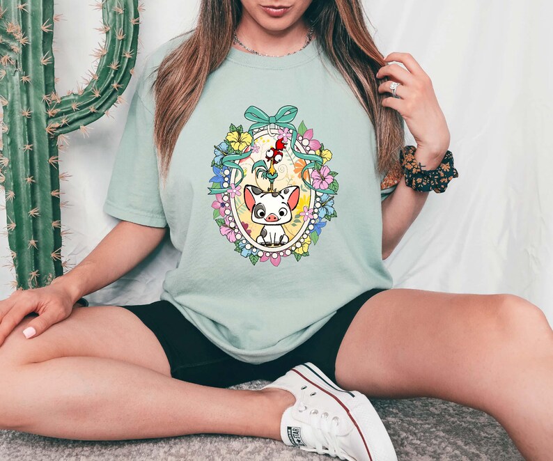 Puede incluir: Camiseta verde claro con una ilustraci&oacute;n de dibujos animados de un cerdo y un gallo dentro de un marco ovalado floral. El dise&ntilde;o incluye un lazo azul y flores coloridas. La camiseta es de corte holgado.