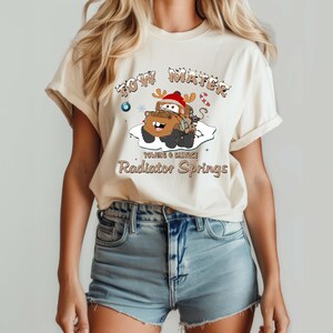 May include: Cream-colored t-shirt featuring a cartoon image of Tow Mater from the movie Cars, wearing a Santa hat and reindeer antlers. The text reads "Tow Mater" above the image, and "Towing & Salvage, Radiator Springs" below.