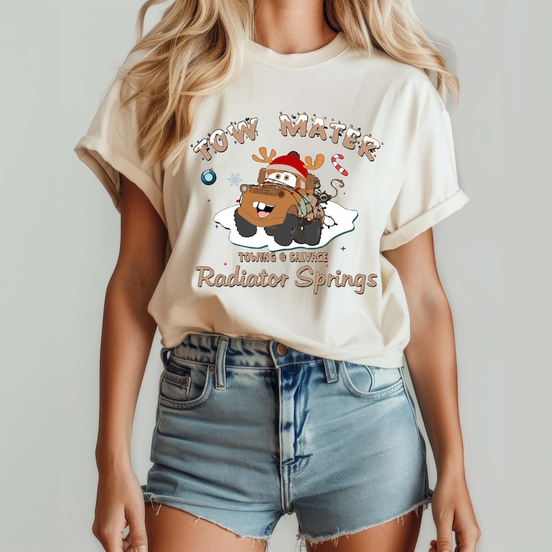 May include: Cream-colored t-shirt featuring a cartoon image of Tow Mater from the movie Cars, wearing a Santa hat and reindeer antlers. The text reads "Tow Mater" above the image, and "Towing & Salvage, Radiator Springs" below.
