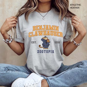 Puede incluir: Camiseta gris jaspeado con la inscripci&oacute;n "BENJAMIN CLAWHAUSER" en naranja y amarillo, y una imagen de dibujos animados de un gato con uniforme de polic&iacute;a. La camiseta tambi&eacute;n incluye el texto "EST. 2016" y "ZOOTOPIA".