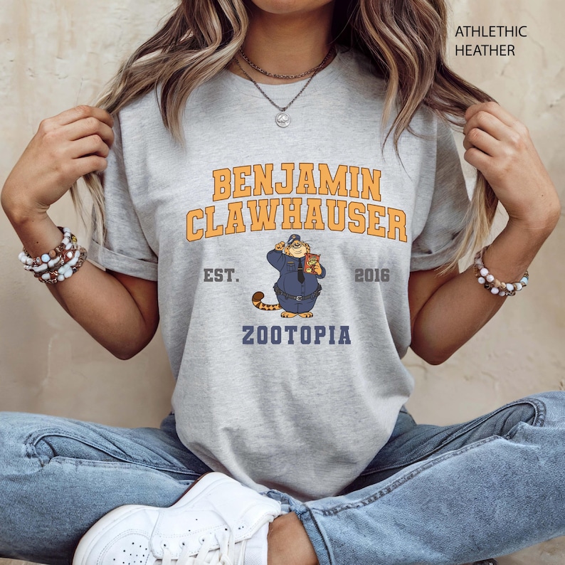 Puede incluir: Camiseta gris jaspeado con la inscripci&oacute;n "BENJAMIN CLAWHAUSER" en naranja y amarillo, y una imagen de dibujos animados de un gato con uniforme de polic&iacute;a. La camiseta tambi&eacute;n incluye el texto "EST. 2016" y "ZOOTOPIA".