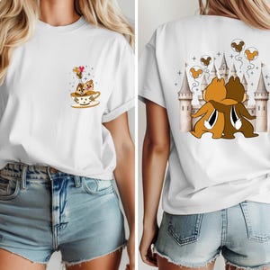 May include: White t-shirt featuring a cartoon illustration of two chipmunks hugging in front of a castle on the back. The front of the shirt has a smaller graphic of the chipmunks in a teacup with balloons. A fun, whimsical design.