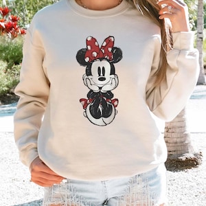 May include: Beige sweatshirt featuring a Minnie Mouse graphic. The graphic depicts Minnie Mouse in a seated pose, with a red bow with white polka dots. The sweatshirt has a crew neck and long sleeves.
