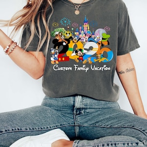 May include: Dark gray t-shirt featuring a colorful graphic with the numbers "2023" and various Disney characters. The text "Custom Family Vacation" is below the numbers. The shirt is worn by a person wearing blue jeans and white shoes.
