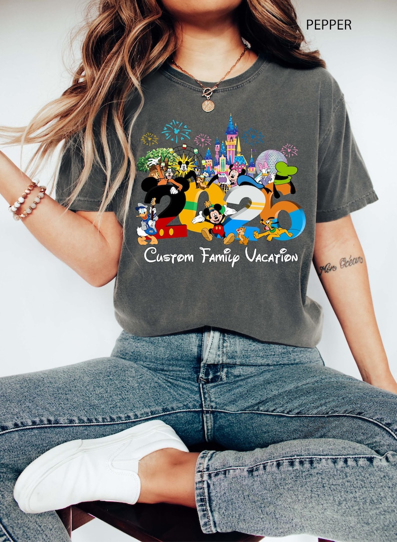 May include: Dark gray t-shirt featuring a colorful graphic with the numbers "2023" and various Disney characters. The text "Custom Family Vacation" is below the numbers. The shirt is worn by a person wearing blue jeans and white shoes.