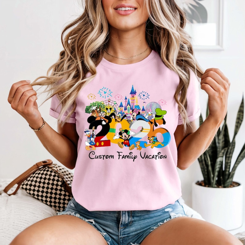 May include: A light pink t-shirt featuring a colorful graphic of Disney characters and the numbers "2026". The text "Custom Family Vacation" is printed below the numbers. The design includes fireworks, a castle, and various cartoon characters.