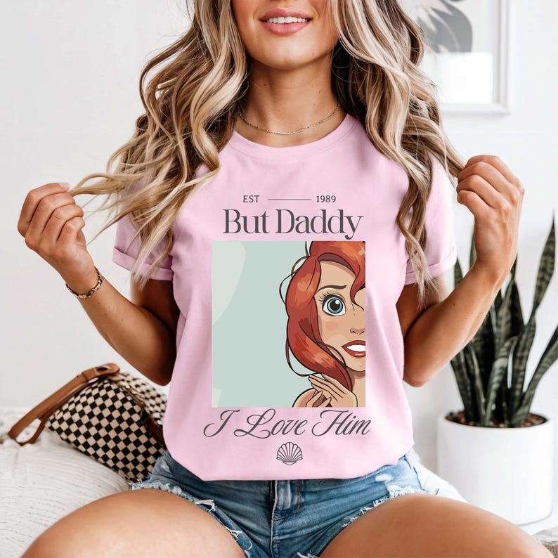 May include: A light pink t-shirt with the text "EST 1989 But Daddy I Love Him" and a graphic of a cartoon character with red hair and blue eyes. The shirt is short-sleeved.