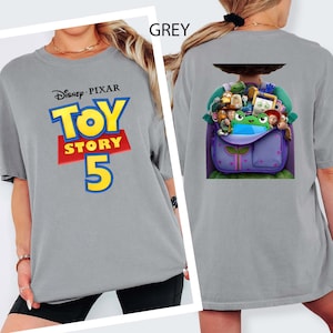 K&ouml;nnte beinhalten: Ein graues T-Shirt mit dem Disney Pixar Toy Story 5 Logo auf der Vorderseite in Blau, Gelb und Rot. Die R&uuml;ckseite des Shirts zeigt eine Grafik eines lila Rucksacks, der mit verschiedenen animierten Spielzeugfiguren gef&uuml;llt ist, darunter eine gr&uuml;ne.