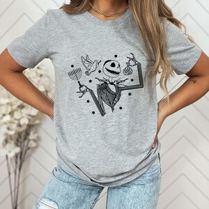 Op de afbeelding: Lichtgrijs t-shirt met een zwarte afbeelding van een skeletpersonage met een menora, een duif en een tas. Het personage draagt een krijtstreep pak. Kleine sterren zijn verspreid rond het ontwerp.