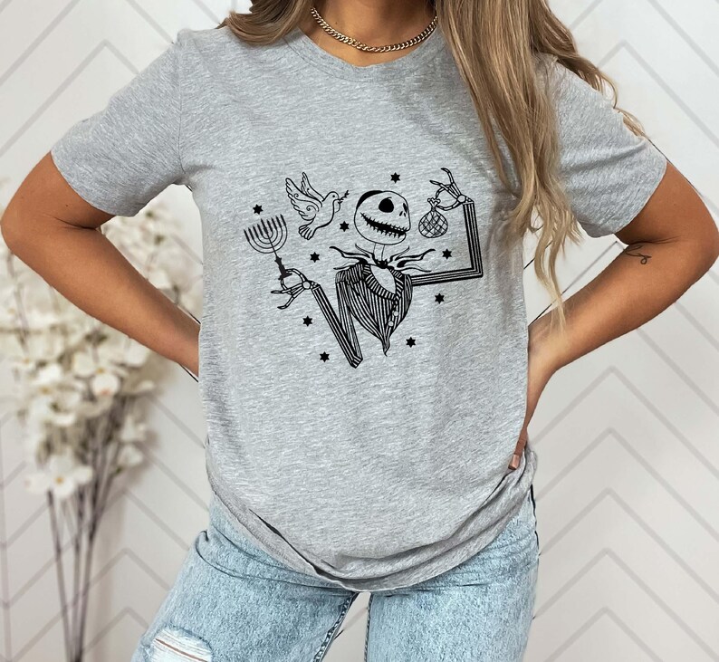 Op de afbeelding: Lichtgrijs t-shirt met een zwarte afbeelding van een skeletpersonage met een menora, een duif en een tas. Het personage draagt een krijtstreep pak. Kleine sterren zijn verspreid rond het ontwerp.