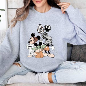 Puede incluir: Una sudadera gris claro con un gr&aacute;fico colorido de Mickey Mouse, Baby Yoda y personajes de Star Wars. El dise&ntilde;o incluye a BB-8, un Stormtrooper y otros elementos ic&oacute;nicos de la franquicia.