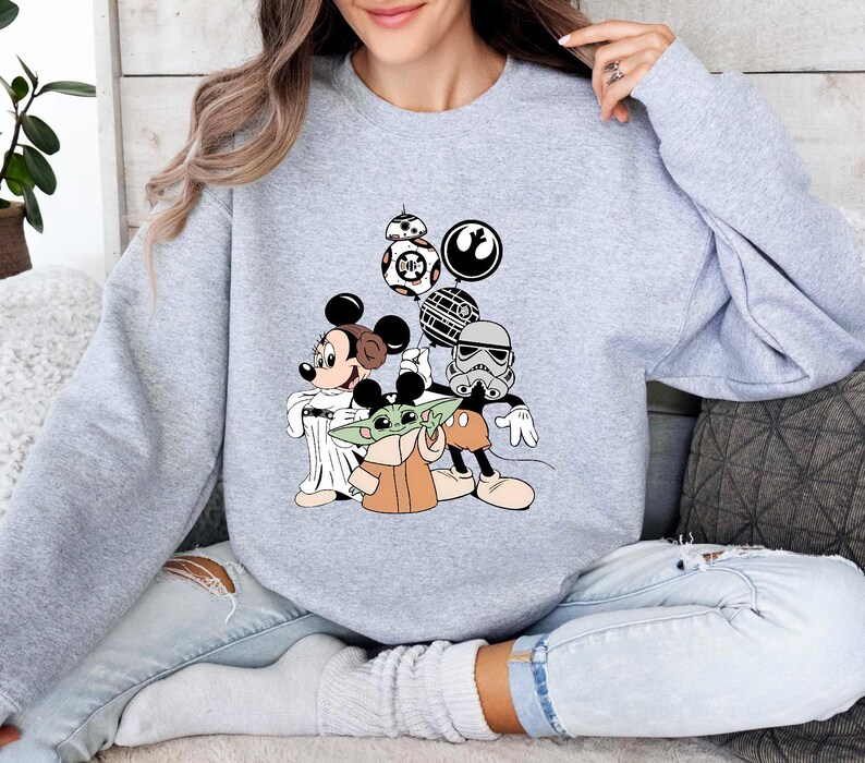 Puede incluir: Una sudadera gris claro con un gr&aacute;fico colorido de Mickey Mouse, Baby Yoda y personajes de Star Wars. El dise&ntilde;o incluye a BB-8, un Stormtrooper y otros elementos ic&oacute;nicos de la franquicia.