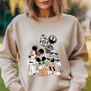 Puede incluir: Sudadera beige con un dise&ntilde;o de Mickey y Minnie Mouse, Baby Yoda y personajes de Star Wars. El dise&ntilde;o incluye a BB-8, un Stormtrooper y otros elementos ic&oacute;nicos de los universos de Star Wars y Disney.