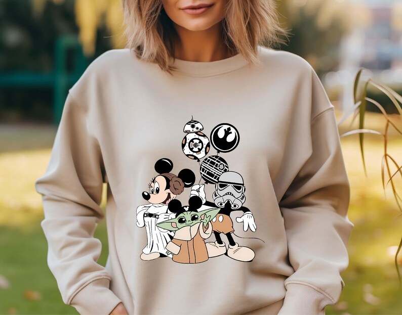 Puede incluir: Sudadera beige con un dise&ntilde;o de Mickey y Minnie Mouse, Baby Yoda y personajes de Star Wars. El dise&ntilde;o incluye a BB-8, un Stormtrooper y otros elementos ic&oacute;nicos de los universos de Star Wars y Disney.
