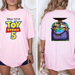 K&ouml;nnte beinhalten: Ein hellrosa T-Shirt mit dem Disney Pixar Toy Story 5 Logo auf der Vorderseite. Die R&uuml;ckseite des Shirts zeigt eine Grafik eines violetten Rucksacks, der mit verschiedenen animierten Spielfiguren gef&uuml;llt ist, darunter eine gr&uuml;ne Froschfigur.