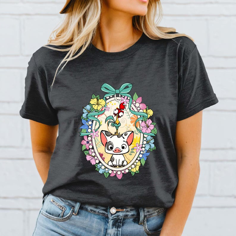 Puede incluir: Camiseta gris oscuro con un dise&ntilde;o ovalado colorido con personajes de dibujos animados. El dise&ntilde;o incluye un cerdo, un gallo y flores. Una cinta turquesa remata el &oacute;valo. La camiseta es de cuello redondo.