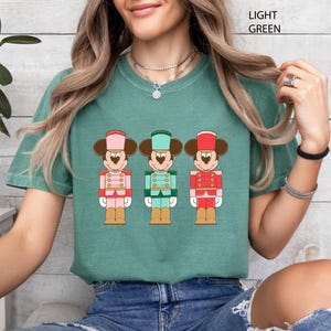 May include: Light green t-shirt featuring a graphic of three Mickey Mouse nutcracker figures in pink, green, and red uniforms. The text "LIGHT GREEN" is printed at the top right of the image. The shirt is worn by a person.