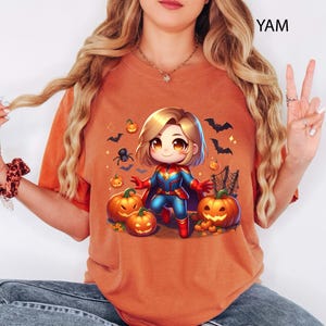 May include: Orange t-shirt featuring a cartoon illustration of a superhero character with blonde hair, wearing a blue and red costume, surrounded by pumpkins, bats, and a spider. The shirt has the text "YAM" in the upper right corner.
