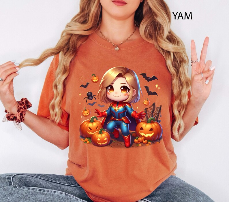 May include: Orange t-shirt featuring a cartoon illustration of a superhero character with blonde hair, wearing a blue and red costume, surrounded by pumpkins, bats, and a spider. The shirt has the text "YAM" in the upper right corner.