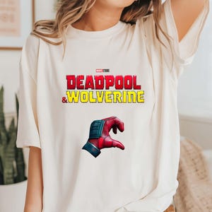 Puede incluir: Camiseta color crema con el texto "DEADPOOL & WOLVERINE" en rojo y amarillo, con un logo de Marvel Studios. Debajo, un gr&aacute;fico de una mano roja y azul.