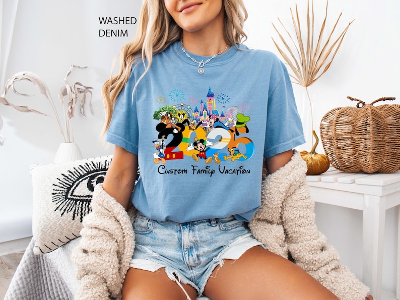 May include: Light blue t-shirt with a colorful graphic featuring Disney characters and the text "2020 Custom Family Vacation." The shirt is made of washed denim material.
