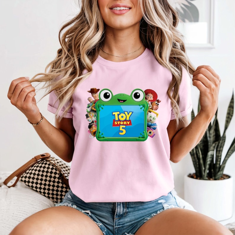 May include: A light pink t-shirt featuring a graphic of a green frog-shaped frame surrounding the text "TOY STORY 5". The frame is flanked by various animated characters including a cowboy, a cowgirl, and other figures from the film franchise.