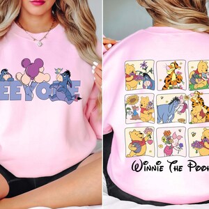 May include: Pink sweatshirt with a design of Eeyore and a balloon on the front. The back of the sweatshirt features a grid of Winnie the Pooh characters. The text "Eeyore" is in blue letters. The text "Winnie The Pooh" is in black script.