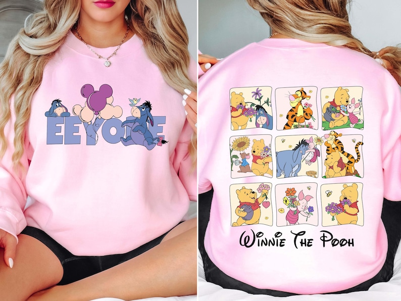 May include: Pink sweatshirt with a design of Eeyore and a balloon on the front. The back of the sweatshirt features a grid of Winnie the Pooh characters. The text "Eeyore" is in blue letters. The text "Winnie The Pooh" is in black script.