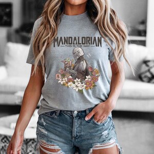 Puede incluir: Camiseta gris con la frase "MANDALORIAN - THIS IS THE WAY" sobre una ilustraci&oacute;n del Mandaloriano y Grogu rodeados de flores. La camiseta es de cuello redondo y manga corta.