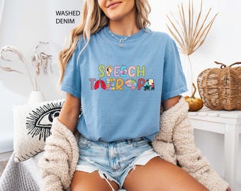 May include: Light blue t-shirt with the words "SPEECH THERAPY" in colorful letters and illustrations of an ear, lungs, brain, and people. The shirt is made of a soft material and is labeled "WASHED DENIM".