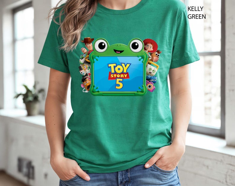 May include: A kelly green short-sleeve t-shirt featuring a graphic of a green frog-shaped frame surrounding the text Toy Story 5. The frame is decorated with various animated character figures including a cowboy, a cowgirl, and a potato-shaped toy.
