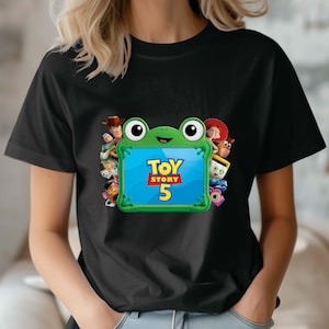 May include: A black t-shirt featuring a central graphic of a green frog-shaped tablet frame. The screen displays the text Toy Story 5 in yellow and red. Surrounding the frog are various animated characters including a cowboy, cowgirl, and potato toy.