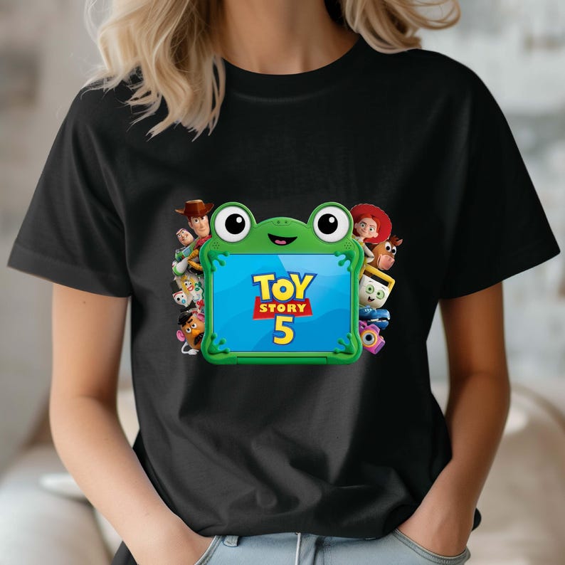 May include: A black t-shirt featuring a central graphic of a green frog-shaped tablet frame. The screen displays the text Toy Story 5 in yellow and red. Surrounding the frog are various animated characters including a cowboy, cowgirl, and potato toy.