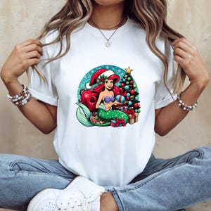 Puede incluir: Camiseta blanca con un dise&ntilde;o de sirena de dibujos animados. La sirena tiene pelo rojo, una cola verde y un gorro de Pap&aacute; Noel. Sostiene una bola de Navidad cerca de un &aacute;rbol decorado y regalos, sobre un fondo azul nevado.