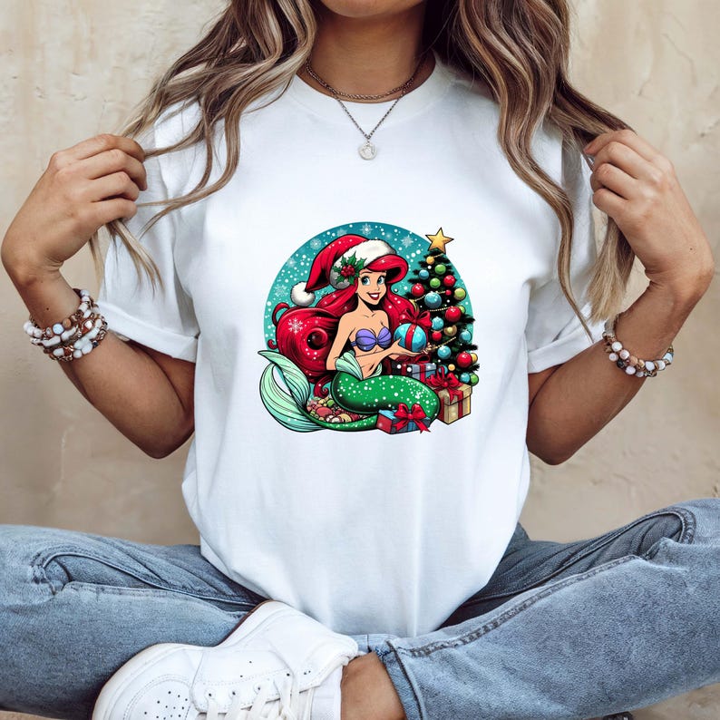 Puede incluir: Camiseta blanca con un dise&ntilde;o de sirena de dibujos animados. La sirena tiene pelo rojo, una cola verde y un gorro de Pap&aacute; Noel. Sostiene una bola de Navidad cerca de un &aacute;rbol decorado y regalos, sobre un fondo azul nevado.