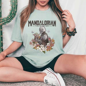 Puede incluir: Camiseta verde claro con la frase "MANDALORIAN - THIS IS THE WAY". La ilustraci&oacute;n muestra al Mandaloriano y a Grogu rodeados de flores y mariposas.